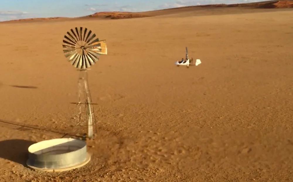 Eine Windpumpe dreht sich in der Namib, dahinter ein Gyrokopter im Flug